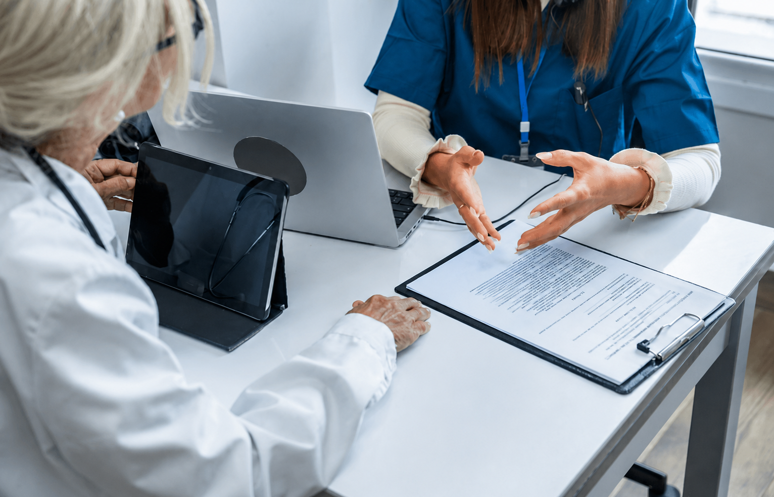 image of two people sitting at a table with a laptop, tablet, and clipboard while discussing.